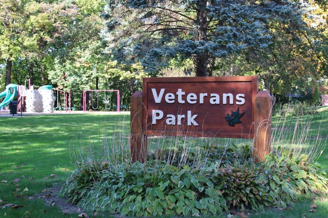 The Veterans Park sign in Appleton, with playground equipment and trees behind it