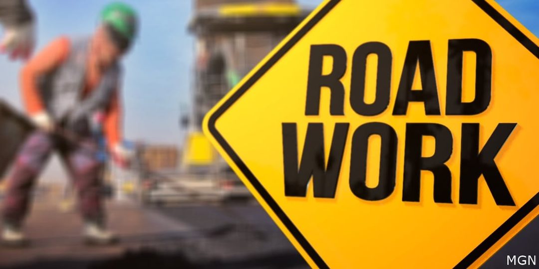 A yellow diamond Road Work sign with a road worker blurred in the background